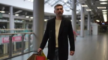 Koyu renk ceketli ve kotlu şık bir adam elinde kırmızı, sarı ve kahverengi alışveriş torbalarıyla modern bir alışveriş merkezinde yürüyor. Gösterişli iç mekan parlak ışıklar, metalik korkuluklar ve...