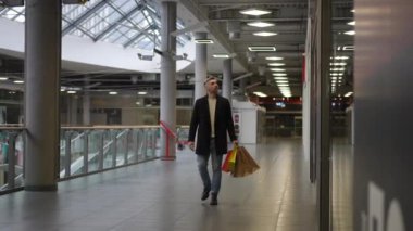 Koyu renk ceketli ve kot pantolonlu şık bir adam elinde renkli alışveriş torbalarıyla modern bir alışveriş merkezinde yürüyor. Geniş iç mekan cam bir tavan, parlak ışıklandırma ve metalik içerir.