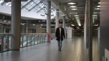 Gündelik giyinmiş ve koyu renk ceketli kendine güvenen bir adam modern bir alışveriş merkezinde yürüyor. Geniş iç mekâna şöyle bir göz atar, camdan bir tavan ve parlak yukarıdan ışıklar görür. Parlak tasarım.