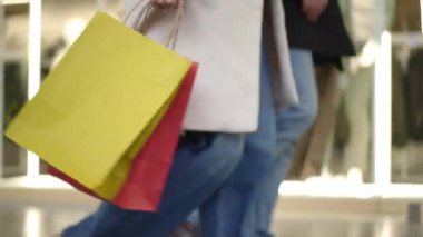 Vitrinlerin önünden geçen şık mankenleri gösteren bir çiftin canlı görüntüsü. Kadın renkli alışveriş torbaları taşırken, erkek de tarafsız tonlu çantalar taşıyarak siyah çantalarını parlaklaştırıyor.