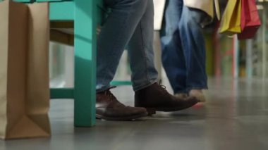 Genç bir adam bir alışveriş merkezindeki turkuaz bir bankta sabırla oturuyor, botları renkli alışveriş torbalarının yanında fayanslı zeminde dinleniyor. Kısa bir süre sonra, bir kadın ek çantalarla gelir, hızla...
