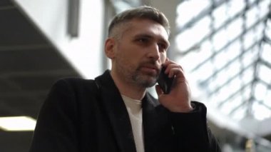 Çağdaş bir ofis binasında yürüyen merdivene tırmanırken akıllı telefonuyla konuşan siyah ceketli profesyonel bir işadamı. Parlak iç mekanda büyük cam pencereler ve geometrik pencereler yer alıyor.