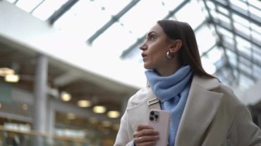 Şık bir kadın modern bir havaalanında akıllı telefonunda kapı yönlerini kontrol ederken yürüyen merdivene çıkıyor. O, bej bir ceket ve açık mavi bir eşarp giyer, pratik ve moda ile harmanlanır. Şey...