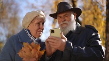 Canlı, renkli bir sonbahar parkında mutlu bir şekilde akıllı telefonu inceleyen yaşlı bir çift neşeli bir anı paylaşıyor.