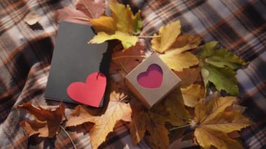 Sevgililer Günü temalı bir hediye kutusunun yakın çekimi. Renkli sonbahar yapraklarıyla çevrili, ekose bir battaniyeye yerleştirilmiş bir kalp tasarımı. Sahnede kırmızı kalpli siyah bir defter var.