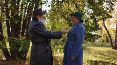 Deri ceketli düşünceli bir adam mavi ceketli ve yeşil şapkalı gülümseyen bir kadına küçük bir buket renkli sonbahar yaprağı sunuyor. Çift güneşli bir parkta hassas bir anı paylaşıyor.