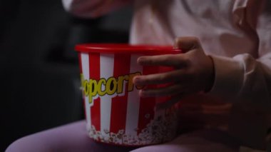 Neşeli bir çocuk heyecanlı bir şekilde büyük çizgili patlamış mısır kovasına uzanıyor, istekli ve film izlemeye hazır.