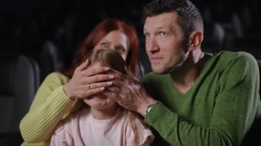 Ebeveynler küçük kızlarının gözlerini kapayıp film hakkında onunla alay ederken aile sinemada neşeli bir anın tadını çıkarıyor. Kız gülümsüyor, ellerini itmeye çalışıyor.