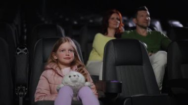 Karanlık sinemada, genç kız gergin bir film sahnesinde gözlerini korkuyla kapar. Bazı izleyiciler arka planda kendini korumaya çalışırken sakince izleyebilirler.