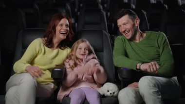 Aile sinemada komedi izlerken keyifli bir anı paylaşıyor. Birlikte gülüyorlar, anne kızını kucaklarken baba mutlu bir şekilde gülümsüyor. Atmosfer dolu.