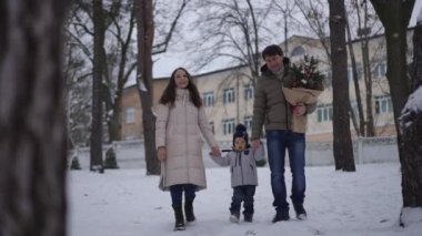 Mutlu bir aile, büyüleyici kış güzelliğinin ortasında bir Noel ağacı taşıyarak karlı bir parkta geziniyor.