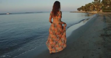 Gün doğumunda kıyıda yürüyen bir kadın, deniz kenarındaki huzurlu sabahın tadını çıkarırken kollarını uzatıyor. Sakin sular ve hafif ışıklandırma dingin ve rahatlatıcı bir atmosfer yaratır.