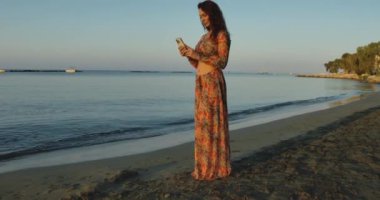 Huzurlu bir an, gün batımında kumsalda çiçekli bir elbise giymiş, nefes kesici manzaranın tadını çıkaran bir kadını gösterir.