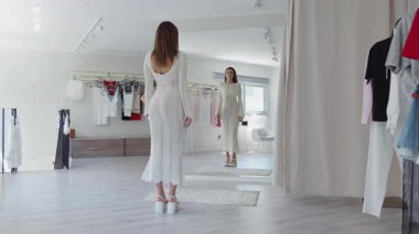 Güzel beyaz bir elbise giymiş zarif bir kadın şık bir butik aynasında yansımasına hayran.