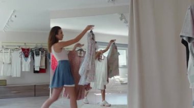 Genç bir kadın, aynadaki tarzına hayranlık duyarken çeşitli kıyafetleri coşkuyla tutuyor.