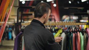 Saçları geriye taranmış, küpe takan şık bir LGBT adamı sarı aksanlı yeşil bir kazak tutuyor ve elbise dükkanına sığıyor mu diye bakıyor. Kişiliği ifade etmek ve moda tercihlerini keşfetmek.