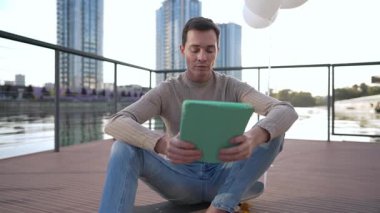 Rıhtımda kaykayının üzerinde oturan genç bir adam, tabletiyle canlı ve modern bir şehirde meşgul.