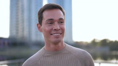 Neşeli bir adam, çevresi modern binalarla çevrili, açık havanın keyfini çıkarırken gülümsüyor.