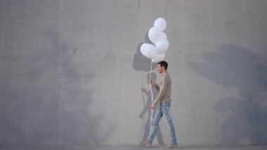 Yalnız bir figür, bir avuç beyaz balonu, desenli beton zemin üzerine tutarak, sakin bir şekilde geziniyor.