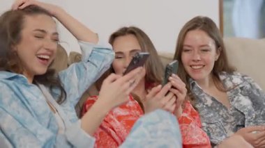Üç yakın arkadaş birlikte rahat bir kanepede akıllı telefonlarını kullanırken neşe ve kahkaha anlarını paylaşıyorlar.