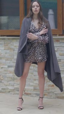 Uzun dalgalı kahverengi saçlı, uzun boylu genç bir kadın leopar desenli bir şalter ve gri bir şalgam içinde duruyor. Yüksek topuklularla ahşap pencereli taş bir duvara karşı zarafetle poz veriyor.