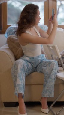 Dalgalı saçlı genç bir kadın başka bir modelin fotoğrafını çekmek için elinde akıllı telefonuyla kanepede oturuyor. Bir çevrimiçi mağaza için moda içeriği yaratmaya odaklanmış durumda. Zarif iç mekan ve stil.