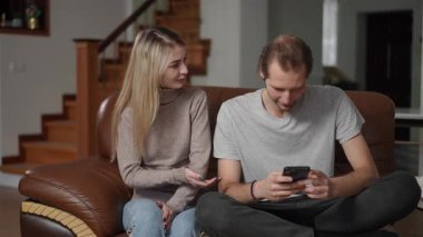 Bir kadın, kocasıyla koltukta oturup telefonuna odaklanarak tartışır. O, elleri ile hayal kırıklığı içinde hareket ederken, o ilgisiz görünüyor. Sahne kapalı bir yerde geçiyor.