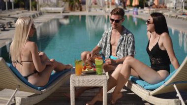 Güneşli bir günde, üç arkadaş havuz kenarında dinlenir, içkilerini yudumlar ve güneşin altında birbirlerinin keyfini çıkarırlar.