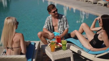 Arkadaşların sıcak güneşin altında içkilerinin ve kahkahalarının tadını çıkardıkları canlı bir havuz kenarı sahnesi.