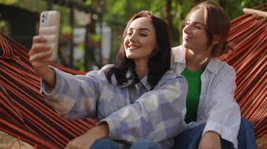 İki yakın arkadaş birlikte selfie çekerken geniş bir gülümseme içinde hamakta keyifli vakit geçiriyorlar.