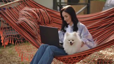 Bir kadın, hamağında tüylü köpeğiyle dizüstü bilgisayar seansından zevk alır. Zamanda huzurlu bir anı yakalar.