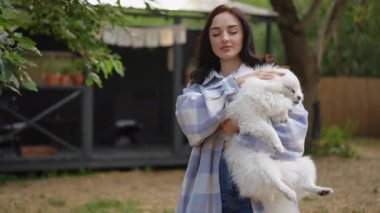 Bir kadın tüylü beyaz bir köpeği nazikçe kucaklıyor, keyifli ve huzurlu bir açık hava anının tadını çıkarıyor.