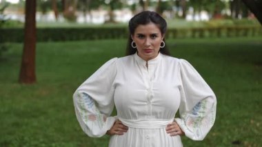 Dalgalı beyaz elbise giyen dengeli bir kadın yemyeşil bir park ortamında kendinden emin bir şekilde duruyor.
