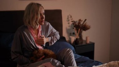 Pijamalı bir kadın rahat yatağında oturuyor, köpeğini nazikçe tutuyor, yatak odasında huzurlu bir atmosfer yaratıyor.