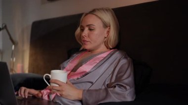 Bir kadın evinde dizüstü bilgisayarı ve sıcak kahvesiyle rahat bir akşamın tadını çıkarır.