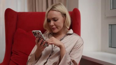 Güzel, ipek bir elbise giymiş bir kadın rahat, davetkar bir yerde akıllı telefonuna bakmaktan mutluluk duyar.