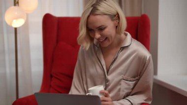 Bir kadın lüks bir kırmızı koltukta elinde bir bardakla otururken rahat ve sıradan bir video görüşmesinden zevk alıyor.