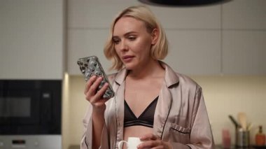 Çekici bir kadın şık mutfağında sabah içkisini yudumlarken akıllı telefonuyla iletişim kuruyor.