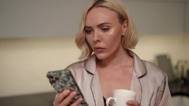 Odaklanmış genç bir kadın, şık bir mutfakta ferahlatıcı bir içkinin tadını çıkarırken akıllı telefonuyla etkileşime girer.