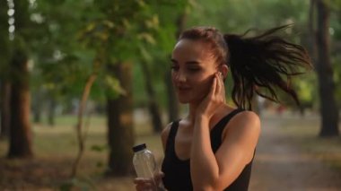 Ağır çekim. Formda genç bir kadın, suyu yudumlarken ve kulaklıklarla müzik dinlerken parkta enerjik bir şekilde koşar.