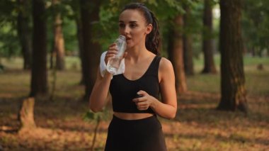 Açık havada egzersiz yapan bir kadın bir an için susuzluğunu gidermek ve huzurlu bir orman ortamında dinlenmek için durur.