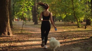Ağır çekim. Bir kadın, tüylü köpeğini gecenin büyüleyici altın saatinde huzurlu bir orman yolunda gezdirir.