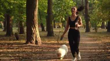 Ağır çekim. Genç bir kadın köpeğiyle birlikte parkta neşeyle koşuyor. Doğada güneşli güzel bir günün tadını çıkarıyor.
