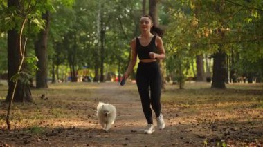 Ağır çekim. Neşeli bir kadın, tüylü köpeğiyle birlikte huzurlu bir parkta koşuyor. Sağlığını ve doğasını tamamen kucaklıyor.