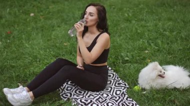 Genç bir kadın, yeşil bir parkta, tüylü beyaz köpeğinin yanında su yudumlayan desenli bir battaniyede oturuyor..