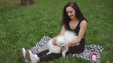 Güneşli güzel bir günde, genç bir kadın parkta oturuyor, rahat bir battaniyede tüylü beyaz köpeğine sarılıyor.