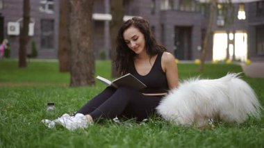 Bir kadın, gür yeşilliklerle çevrili tüylü beyaz köpeğiyle huzurlu bir parkta kitap okumaktan hoşlanır.