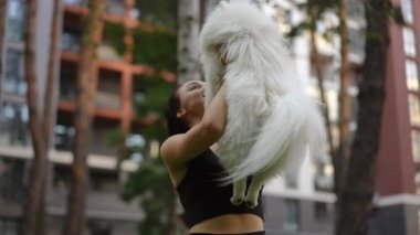 Neşeli bir kadın, tüylü köpeğini güneşli bir parkta sevgisini ve mutluluğunu göstererek kucaklıyor.