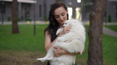 Genç bir kadın, tüylü beyaz bir köpeği şefkatle tutuyor, sakin ve huzurlu bir park ortamının tadını çıkarıyor.