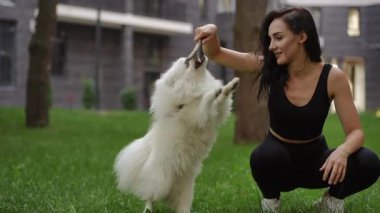 Neşeli bir kadın yemyeşil bir parkta tüylü köpeğiyle neşe içinde etkileşime geçer ve neşeli bağlarını gösterir.